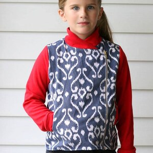 May include: A young person wearing a red long-sleeved shirt with a blue and white patterned jacket over it. The jacket has a zipper down the front and a pocket on the left side. The person is wearing blue jeans.