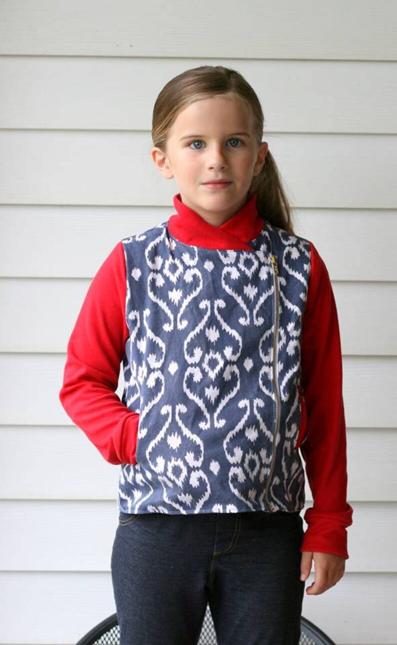 May include: A young person wearing a red long-sleeved shirt with a blue and white patterned jacket over it. The jacket has a zipper down the front and a pocket on the left side. The person is wearing blue jeans.