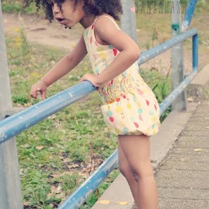 Puede incluir: Una ni&ntilde;a peque&ntilde;a con el pelo rizado marr&oacute;n lleva un vestido estampado de colores con un dobladillo con volantes. Est&aacute; de pie sobre una barandilla de metal azul y sonr&iacute;e.