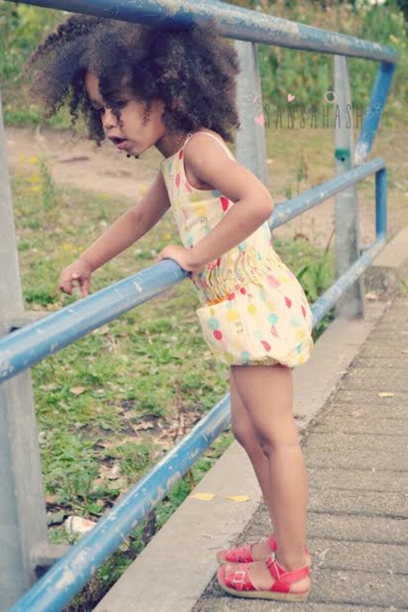 Puede incluir: Una ni&ntilde;a peque&ntilde;a con el pelo rizado marr&oacute;n lleva un vestido estampado de colores con un dobladillo con volantes. Est&aacute; de pie sobre una barandilla de metal azul y sonr&iacute;e.