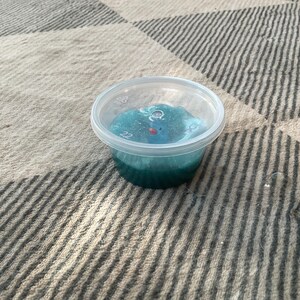 以下が含まれることがあります： 青緑色のスライムが入った小さな透明なプラスチック容器。スライムは層状の外観で、底は濃い色合いです。容器は模様のある布の上に置かれています。
