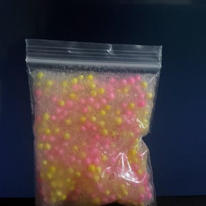 May include: A clear plastic bag filled with small, round beads. The beads are a mix of pink and yellow, creating a colorful and textured appearance. The bag is sealed at the top.