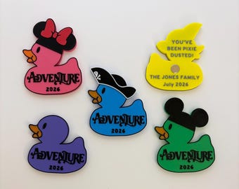 Puede incluir: Cinco imanes planos de goma con forma de pato. Uno rosa con lazo, uno azul con sombrero pirata, uno morado, uno verde con orejas de ratón y uno amarillo con el texto YOU'VE BEEN PIXIE DUSTED! THE JONES FAMILY July 2026. Otros muestran ADVENTURE 2026.