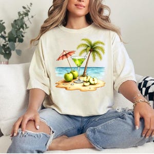 May include: A cream-colored short-sleeve t-shirt featuring a graphic print of a tropical beach scene. The design includes a green cocktail in a glass, a palm tree, a red and white striped umbrella, and several green apples on the sand.