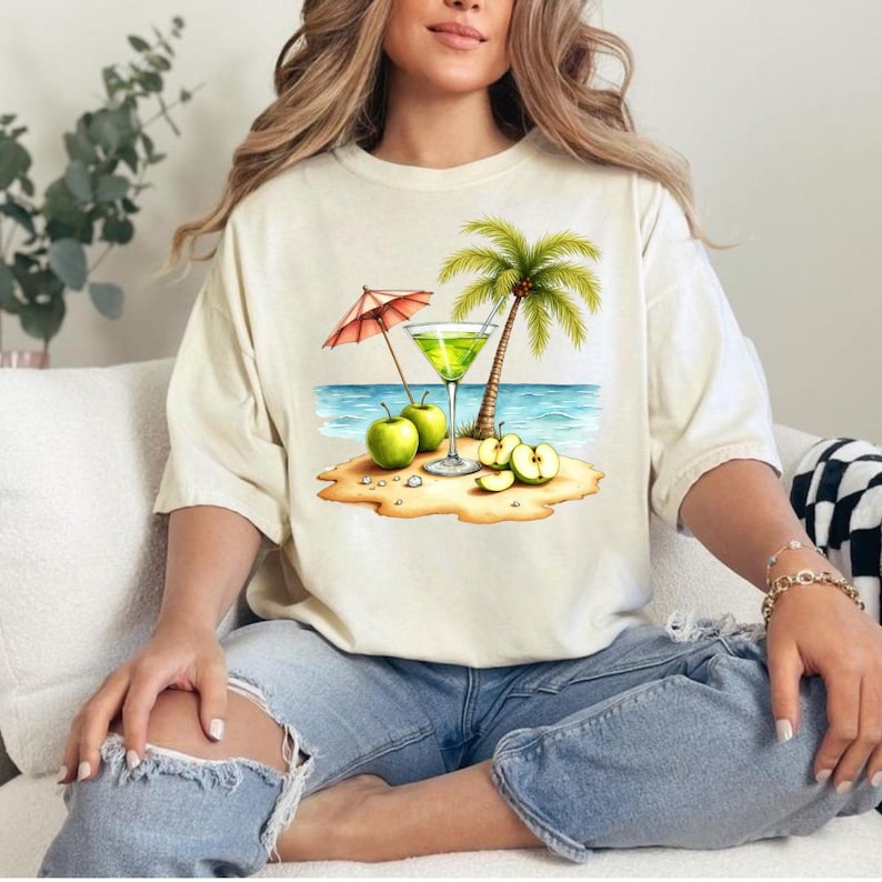 May include: A cream-colored short-sleeve t-shirt featuring a graphic print of a tropical beach scene. The design includes a green cocktail in a glass, a palm tree, a red and white striped umbrella, and several green apples on the sand.