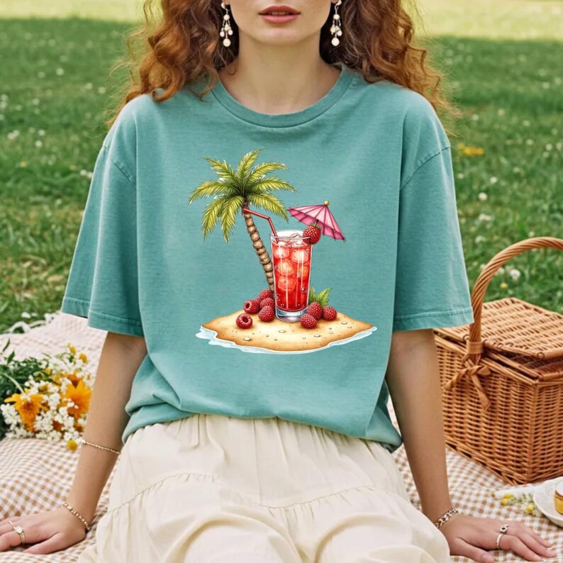May include: A sage green short-sleeve t-shirt featuring a graphic print of a tropical island scene. The design includes a palm tree, a tall glass of red fruit punch with a pink umbrella, and several fresh raspberries on a small sandy patch.
