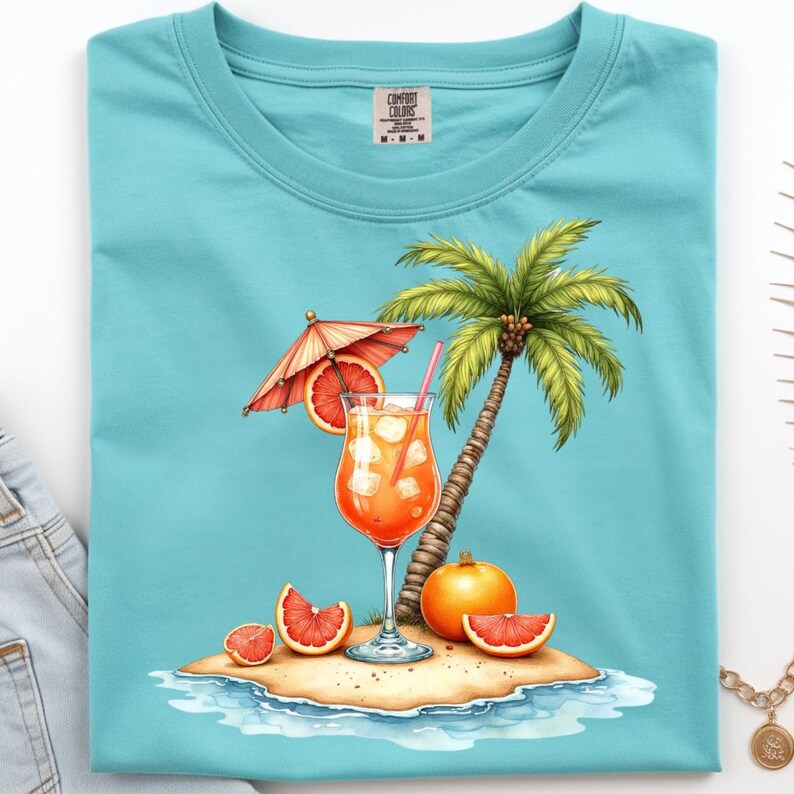 May include: A turquoise t-shirt featuring a graphic of a tropical island scene with a palm tree, a glass of orange cocktail with ice and a pink straw, a grapefruit slice, a small umbrella, and a whole orange on a sandy beach with water.