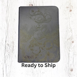 May include: A rectangular gray trading card binder featuring a subtle, monochromatic embossed design of three water-type creatures in various poses. The lower left corner displays the word vaultx in a clean, sans-serif font on the cover.