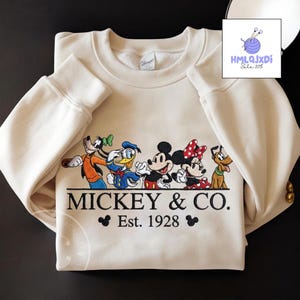 A white sweatshirt with a graphic design featuring Disney characters Mickey Mouse and Donald Duck, along with the text "MICKEY & CO. Est. 1928".