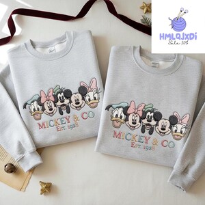 two gray sweatshirts with a group of Disney characters, including Mickey Mouse, Donald Duck, Goofy, and Minnie Mouse, printed on them. The sweatshirts are displayed on a white surface, with a red ribbon and some decorations in the background.
