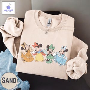 A beige sweatshirt with a graphic of four Disney Minnie Mouse characters, including Snow White and the Seven Dwarfs, printed on it.