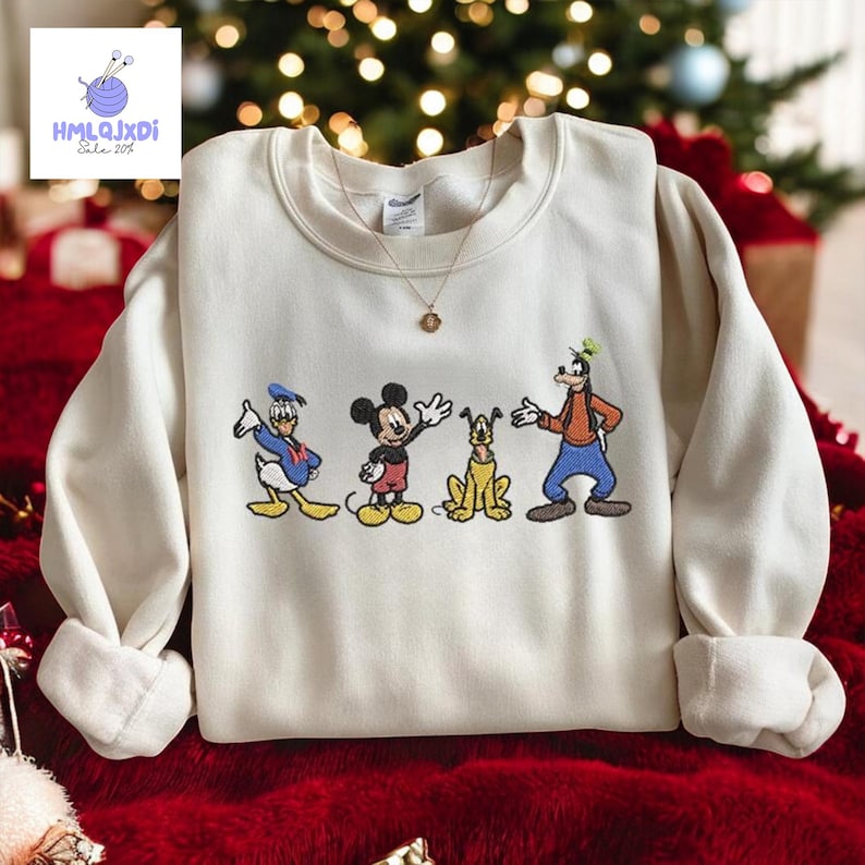 A white sweater with a graphic of Disney characters Mickey Mouse, Donald Duck, and Goofy on it, along with a gold necklace. The sweater is displayed on a red blanket, and there is a Christmas tree in the background.