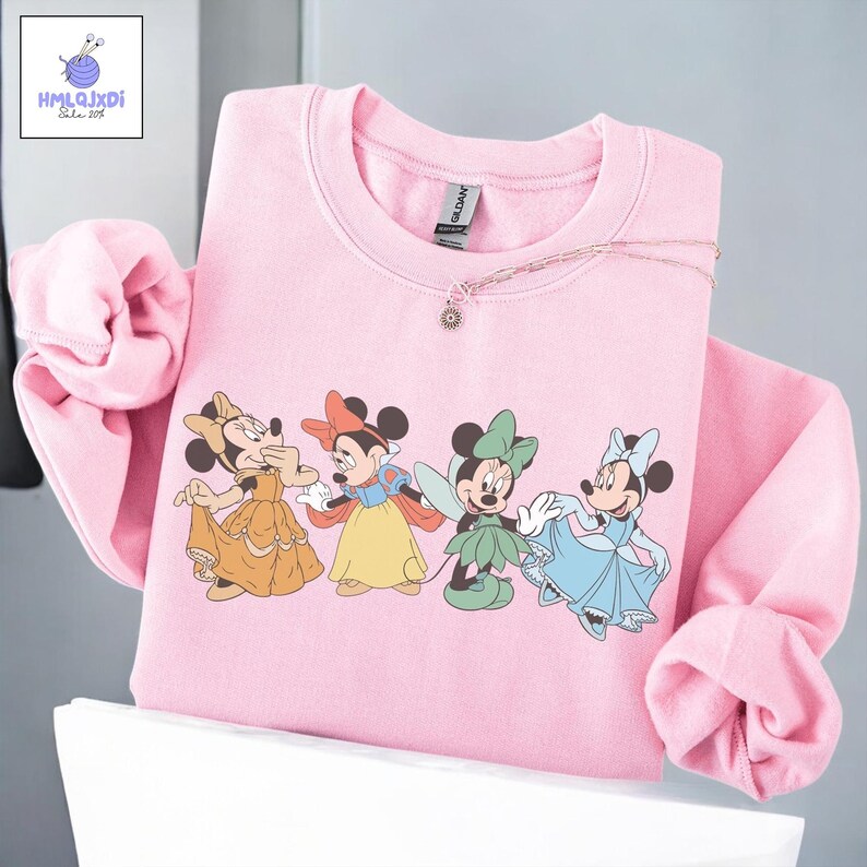A pink sweatshirt with a graphic of four Disney Minnie Mouse characters, including Snow White, Cinderella, Paddington Bear, and Mary Poppins, printed on the front.