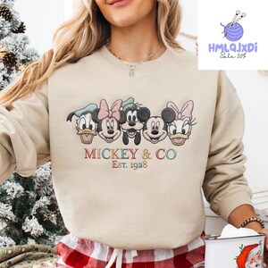 A woman wearing a beige sweater with a graphic of Mickey Mouse and Minnie Mouse, along with the text "Mickey & Co Est. 1928".