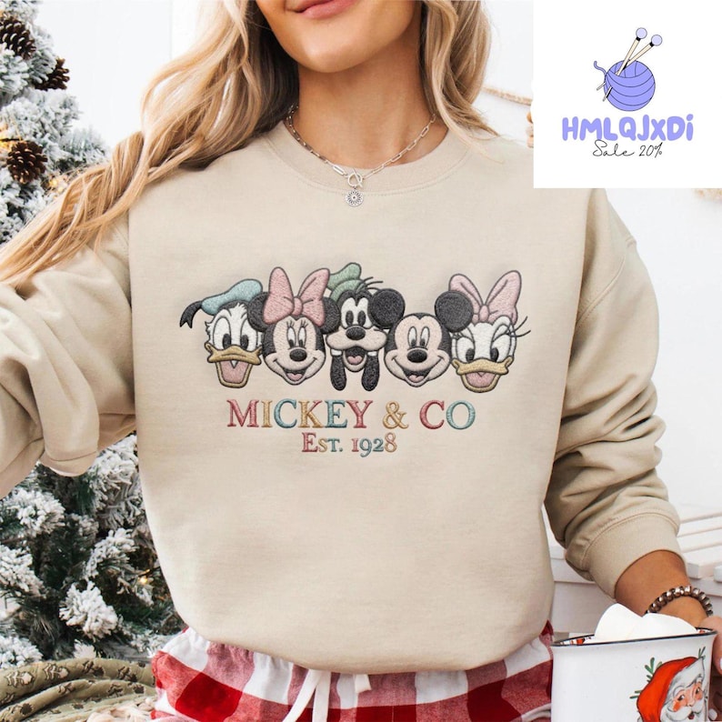 A woman wearing a beige sweater with a graphic of Mickey Mouse and Minnie Mouse, along with the text "Mickey & Co Est. 1928".