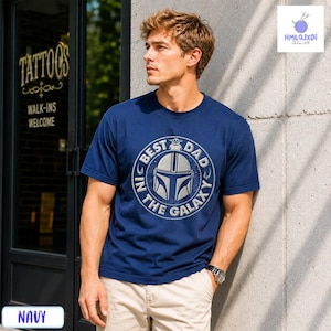A man wearing a navy blue t-shirt with a graphic design stands in front of a tattoo shop.