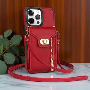 以下が含まれることがあります： 赤い革の携帯電話ケース、クロスボディストラップ付き。ケースには、金色のジッパーと留め金が付いた小さな財布が付いています。この携帯電話ケースはスマートフォンを収納できるように設計されており、黒い縁があります。