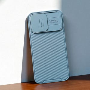 May include: A light blue phone case with a sliding camera cover. The case has a textured, diagonal striped pattern and the words "Camera Protection" are printed on the cover. The case is leaning against a white wall and resting on a wooden surface.