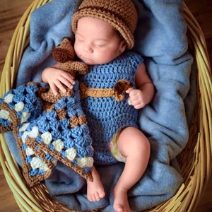 Pu&ograve; includere: Un neonato con un vestito di uncinetto blu e un cappello di uncinetto marrone sta dormendo in un cesto di vimini. Il bambino tiene un giocattolo di uncinetto marrone e blu.