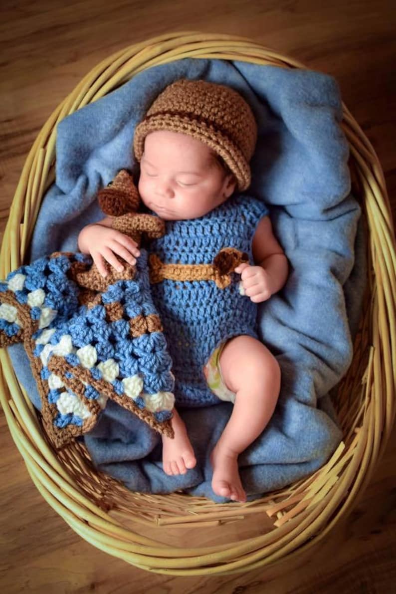 Pu&ograve; includere: Un neonato con un vestito di uncinetto blu e un cappello di uncinetto marrone sta dormendo in un cesto di vimini. Il bambino tiene un giocattolo di uncinetto marrone e blu.