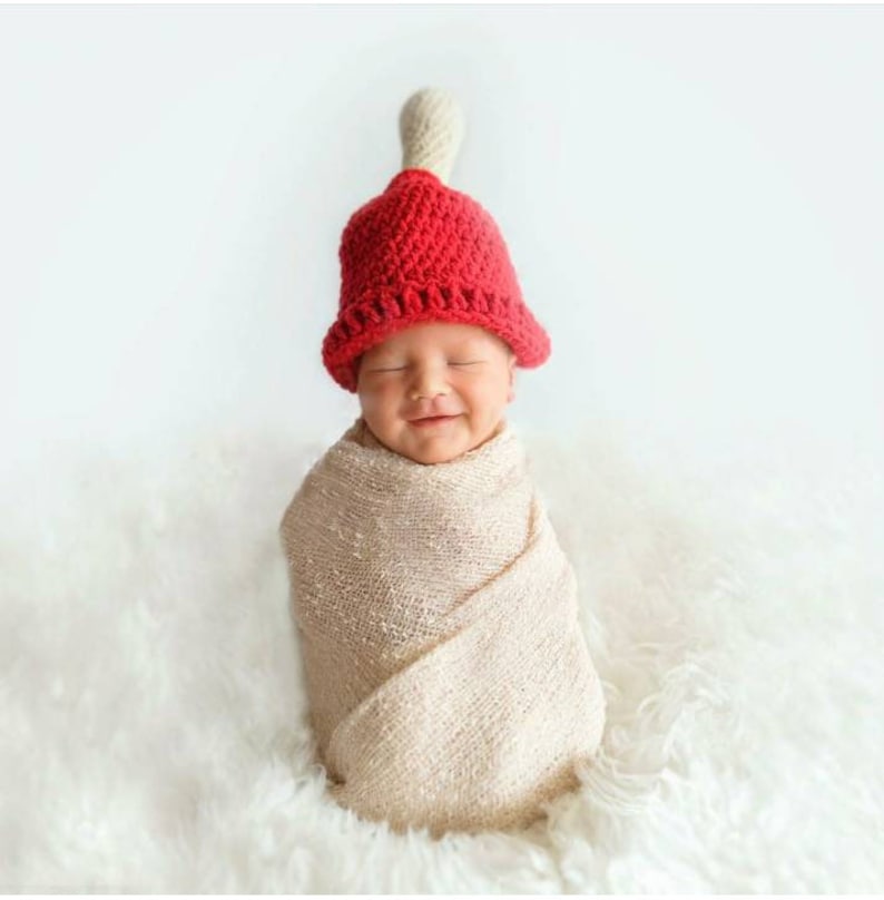 Puede incluir: Un beb&eacute; con un gorro rojo de campana de ganchillo con un pomp&oacute;n blanco en la parte superior, envuelto en una manta beige, sonriendo a la c&aacute;mara.