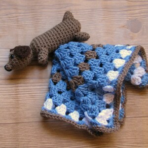 Pu&ograve; includere: Un giocattolo di dachshund di uncinetto marrone su una coperta di uncinetto blu e bianca.