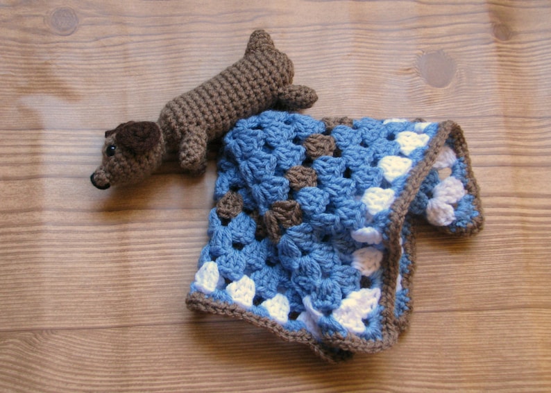 Pu&ograve; includere: Un giocattolo di dachshund di uncinetto marrone su una coperta di uncinetto blu e bianca.