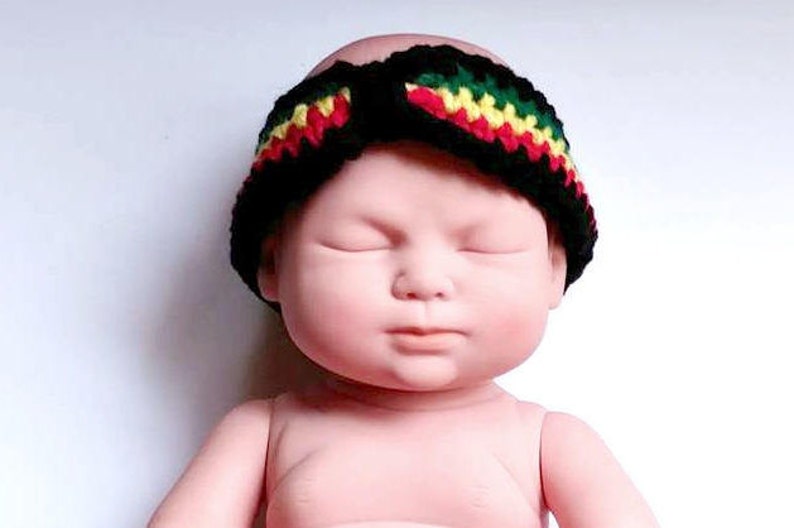 May include: A black crochet headband with red, yellow, and green stripes. The headband is worn by a doll. The headband is a baby accessory.