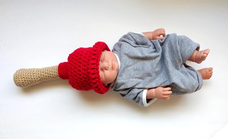 Puede incluir: Un gorro de punto rojo con un mango de punto beige, que se asemeja a un pincel, es usado por un beb&eacute; de juguete acostado sobre una superficie blanca.