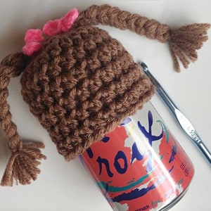 May include: A brown crocheted hat with two brown braids and pink bows. The hat is sitting on top of a can of sparkling water. A crochet hook is lying on the table next to the hat.