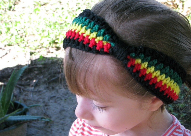 May include: A black crochet headband with green, yellow, and red stripes. The headband is worn on a person's head. The colors are arranged in a pattern, creating a vibrant and eye-catching accessory. The headband is a handmade item.