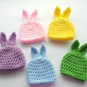 May include: Five crocheted bunny hats in different colors: pink, purple, light blue, yellow, and green. The hats are all made with a similar stitch pattern and have long, floppy ears.