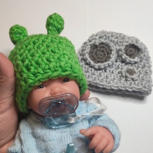 May include: A green crocheted hat with two small bumps on top, a gray crocheted hat with two round holes, and a baby doll wearing a blue shirt and a pacifier.