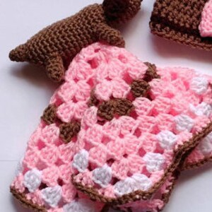 Pu&ograve; includere: Una copertina per neonati all'uncinetto con uno schema di colori rosa, bianco e marrone. &Egrave; incluso un piccolo cane all'uncinetto marrone.