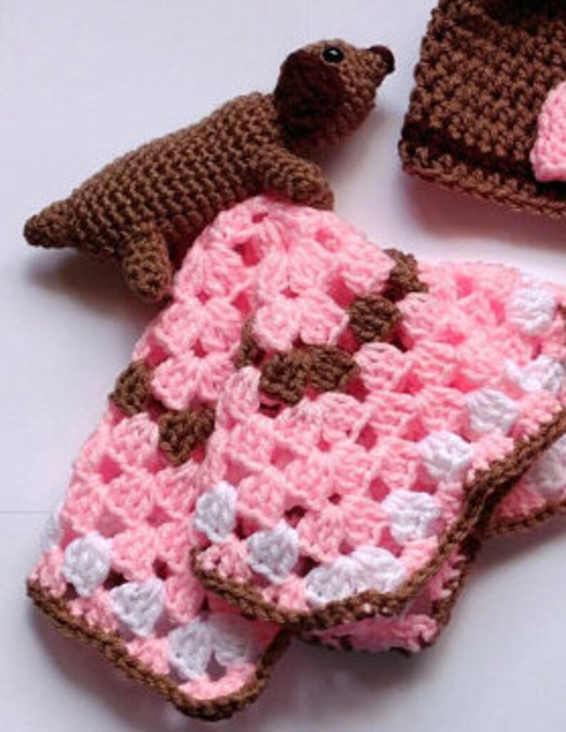 Pu&ograve; includere: Una copertina per neonati all'uncinetto con uno schema di colori rosa, bianco e marrone. &Egrave; incluso un piccolo cane all'uncinetto marrone.