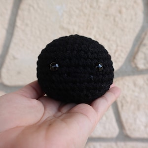May include: A small, black crocheted ball with two black button eyes. The ball is being held in a person's hand.
