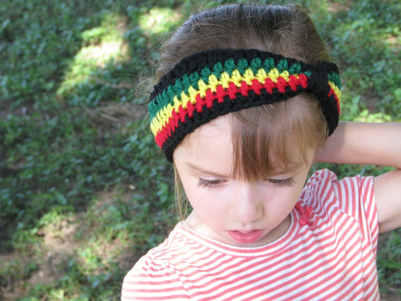 May include: A black, red, yellow, and green crochet headband. The headband is worn by a person wearing a red and white striped shirt. The headband is a vibrant accessory.