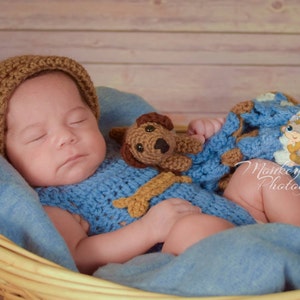 Pu&ograve; includere: Un bambino che dorme con un cappello di uncinetto marrone e un vestito di uncinetto blu con un giocattolo di uncinetto marrone a forma di cucciolo. Il bambino &egrave; sdraiato in un cesto di vimini con una coperta blu.
