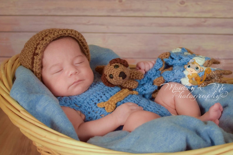 Pu&ograve; includere: Un bambino che dorme con un cappello di uncinetto marrone e un vestito di uncinetto blu con un giocattolo di uncinetto marrone a forma di cucciolo. Il bambino &egrave; sdraiato in un cesto di vimini con una coperta blu.