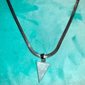 May include: A silver-toned necklace with a triangular pendant. The pendant is a light, translucent color, and the chain has a woven design. The necklace is displayed against a teal background.