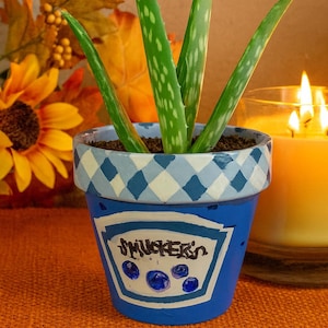 May include: A blue painted flower pot with a white and blue checkered rim, featuring the word "Smuckers" and blueberry illustrations. The pot holds an aloe vera plant with green, spotted leaves. A lit candle and fall foliage are in the background.