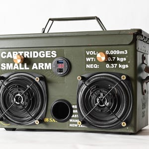 May include: An olive green ammunition box converted into a speaker system. The box has two black speakers with grilles, a digital display, and the text "CARTRIDGES SMALL ARM". It includes a handle and brass-coloured details.