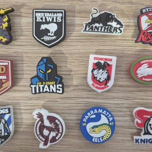 Könnte beinhalten: Eine Sammlung von Logo-Anhängern von Sportmannschaften, darunter "STORM", "NEW ZEALAND KIWIS", "PANTHERS" und "SYDNEY Roosters". Weitere Anhänger zeigen "QLD MAROONS", "GOLD COAST TITANS", "ST GEORGE", "SOUTH SYDNEY RABBITOHS", "BULLDOGS", "PARRAMATTA EELS" und "KNIGHTS".