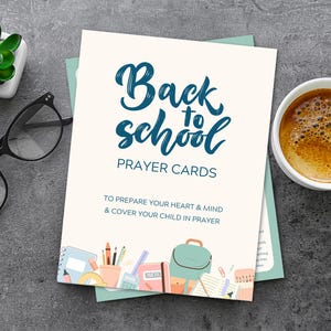 May include: A stack of "Back to School Prayer Cards" with a teal and white design, a cup of coffee, a small succulent, and black glasses on a gray surface. The cards have school-themed illustrations.