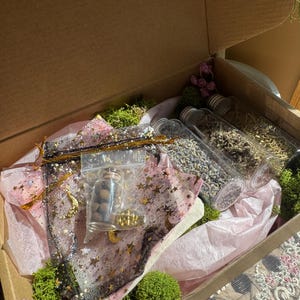 May include: An open cardboard box filled with various items. Inside are small glass jars, a black mesh bag with gold stars, pink tissue paper, green moss, and dried flowers. The box is lit by sunlight.