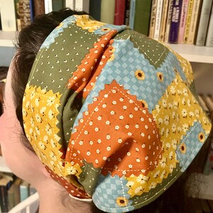 May include: A patchwork fabric headband featuring a mix of floral and geometric patterns in yellow, orange, green, and blue. The headband is made of stitched fabric squares, creating a quilted effect. The design includes sunflowers and small white flowers.