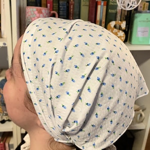 May include: White headscarf with a repeating pattern of small blue flowers. The fabric is gathered at the top of the head and drapes down the sides. The head covering is likely made of a lightweight material.