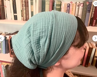 Op de afbeelding: Een lichtblauwe hoofdbedekking van een textuurstof. De hoofddoek is over het hoofd gedrapeerd en bedekt het haar. De achtergrond bevat een boekenkast vol boeken.