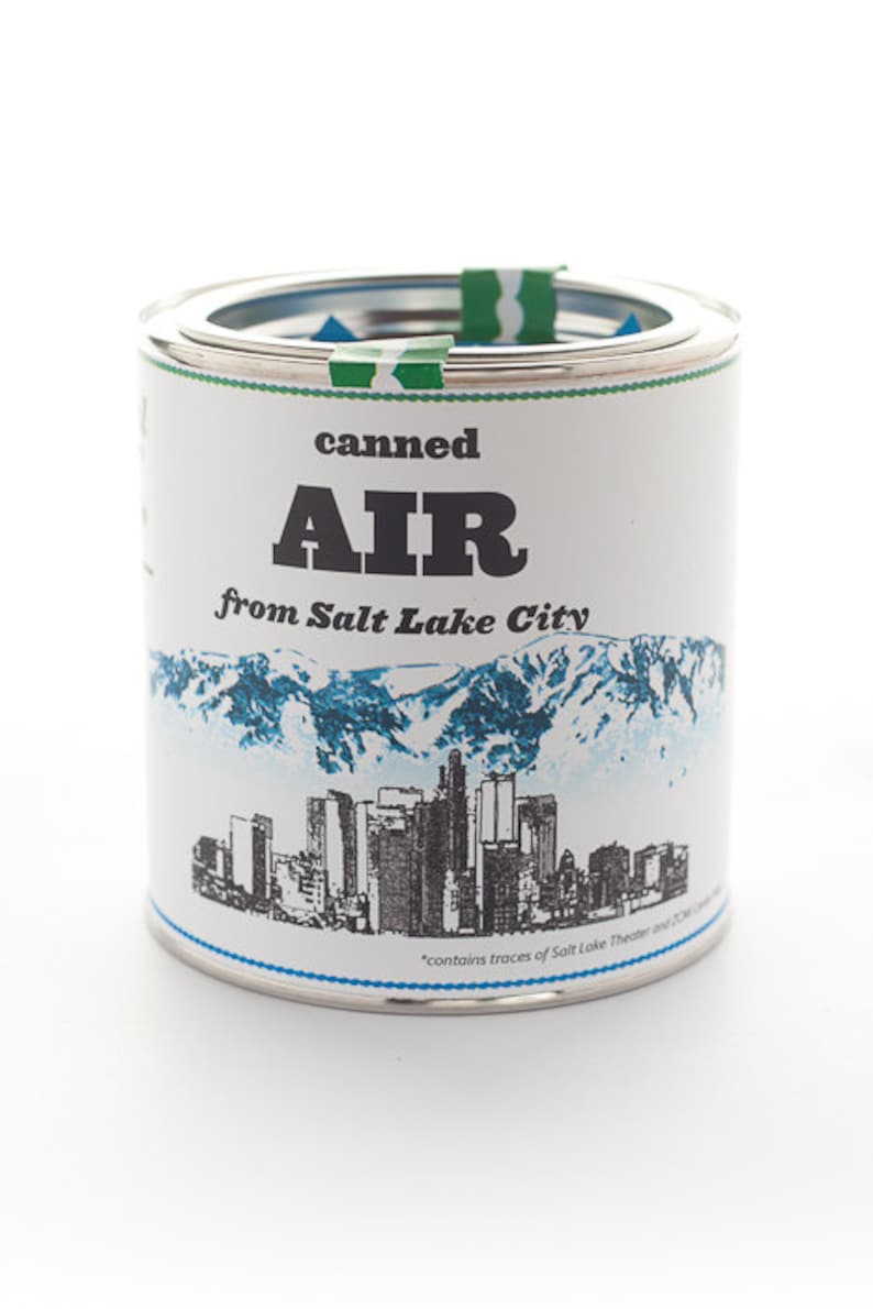 Pu&ograve; includere: Una lattina di metallo bianca con un'immagine blu e bianca di uno skyline cittadino e montagne. La lattina dice "canned AIR from Salt Lake City" e "contains traces of Salt Lake Theater and 2010 Olympics".
