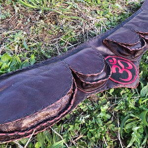May include: A brown leather belt with multiple pockets and a red Om symbol patch. The belt is lying on a bed of green grass.
